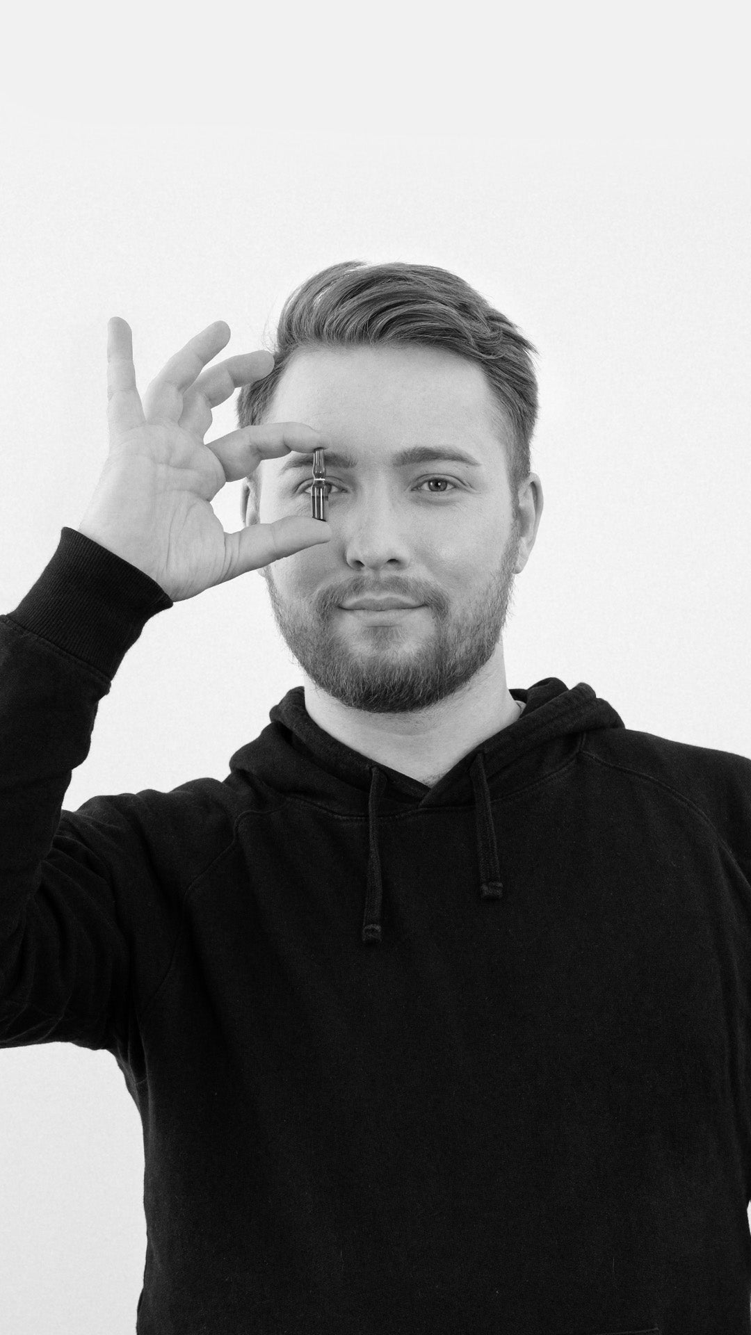 Ein junger Mann hält eine kleine Braunglasflasche vor sein Auge und blickt dabei in die Kamera. Er trägt einen schwarzen