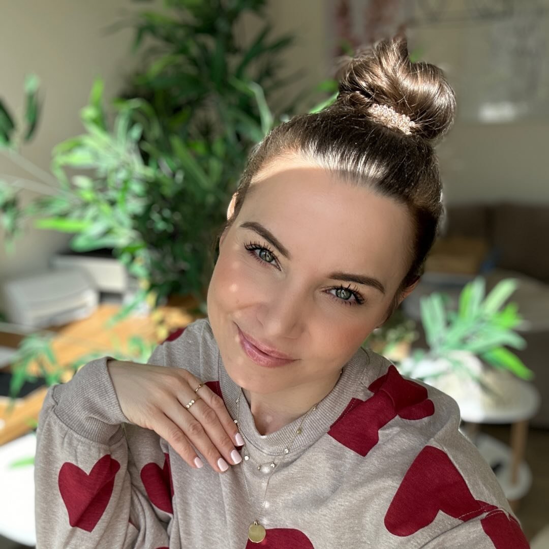 Eine Frau mit einem hohen Dutt und einem lächelnden Gesicht trägt einen Pullover mit Herzmotiven. Im Hintergrund sind