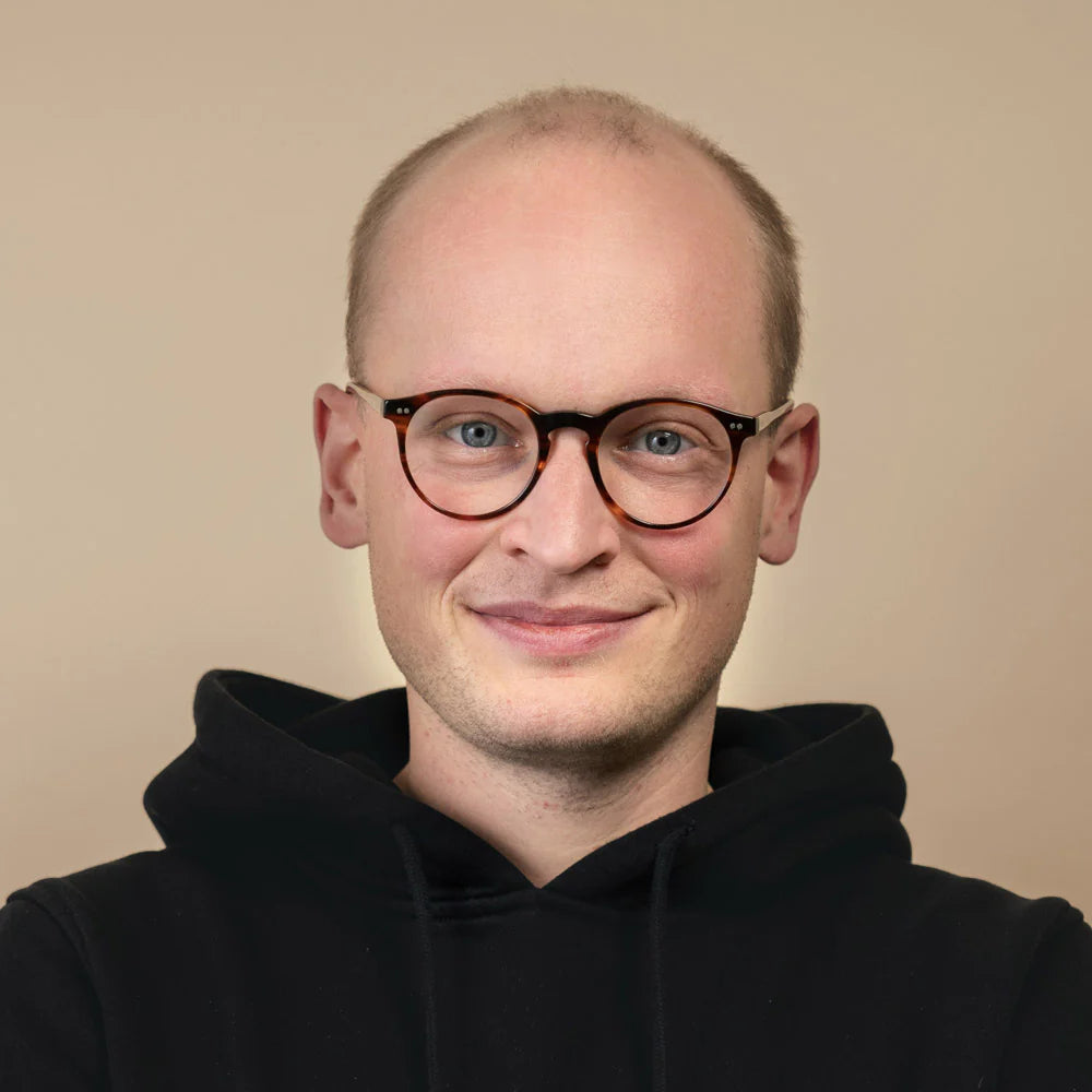 Porträt eines Mannes mit Brille und einem schwarzen Hoodie, vor einem neutralen, hellen Hintergrund.
