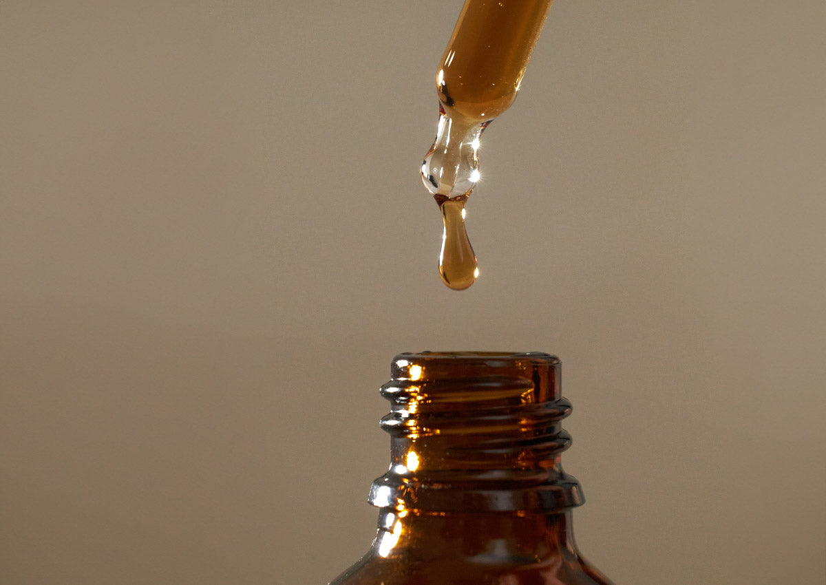 Tropfen einer goldenen Flüssigkeit fallen von einer Pipette in eine braune Glasflasche.