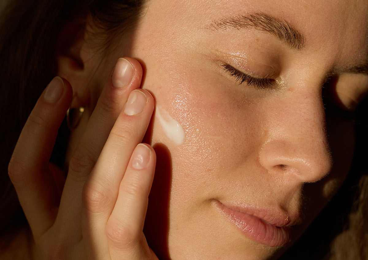 Frau mit geschlossenen Augen trägt eine Creme auf die Wange auf, sanfte Lichtverhältnisse betonen die gesunde Sommerbräune.