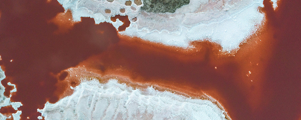 Luftaufnahme eines rötlichen Salzsees mit salzüberzogenem Ufer und geologischen Strukturen.