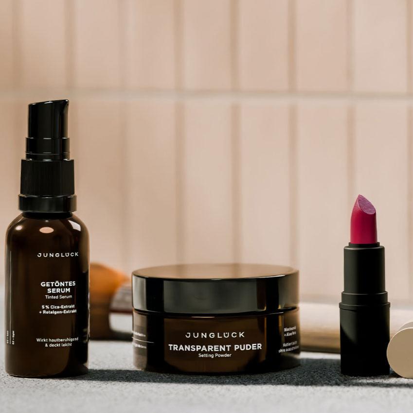 Drei JUNGLÜCK-Produkte auf einem grauen Untergrund: ein Getöntes Serum in einer Braunglasflasche, ein Transparent Puder in