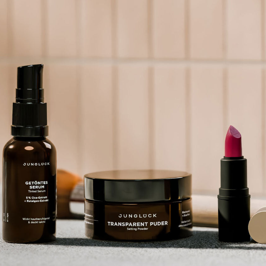 Eine Braunglasflasche mit getöntem Serum, ein schwarzer Tiegel für transparenten Puder und ein Lippenstift in kräftigem