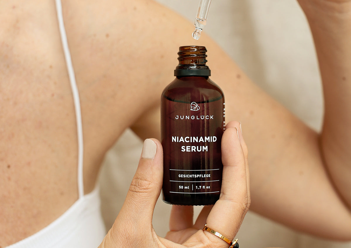 Eine Hand hält eine Braunglasflasche mit Niacinamid Serum. Eine Pipette tropft das Serum auf die Haut.
