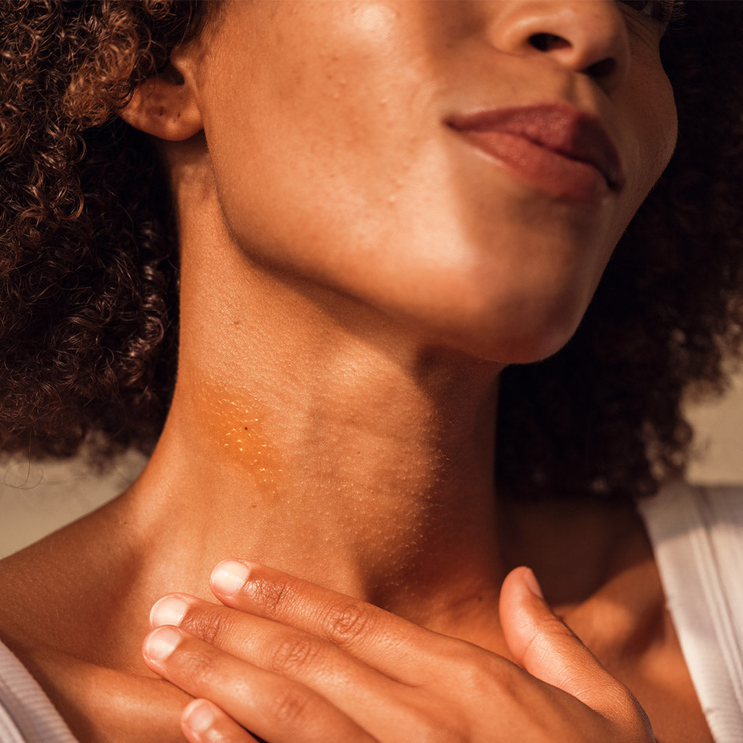 Nahaufnahme einer Frau mit lockigem Haar, die sanft ihren Hals berührt. Ihre Haut wirkt strahlend und gesund.
