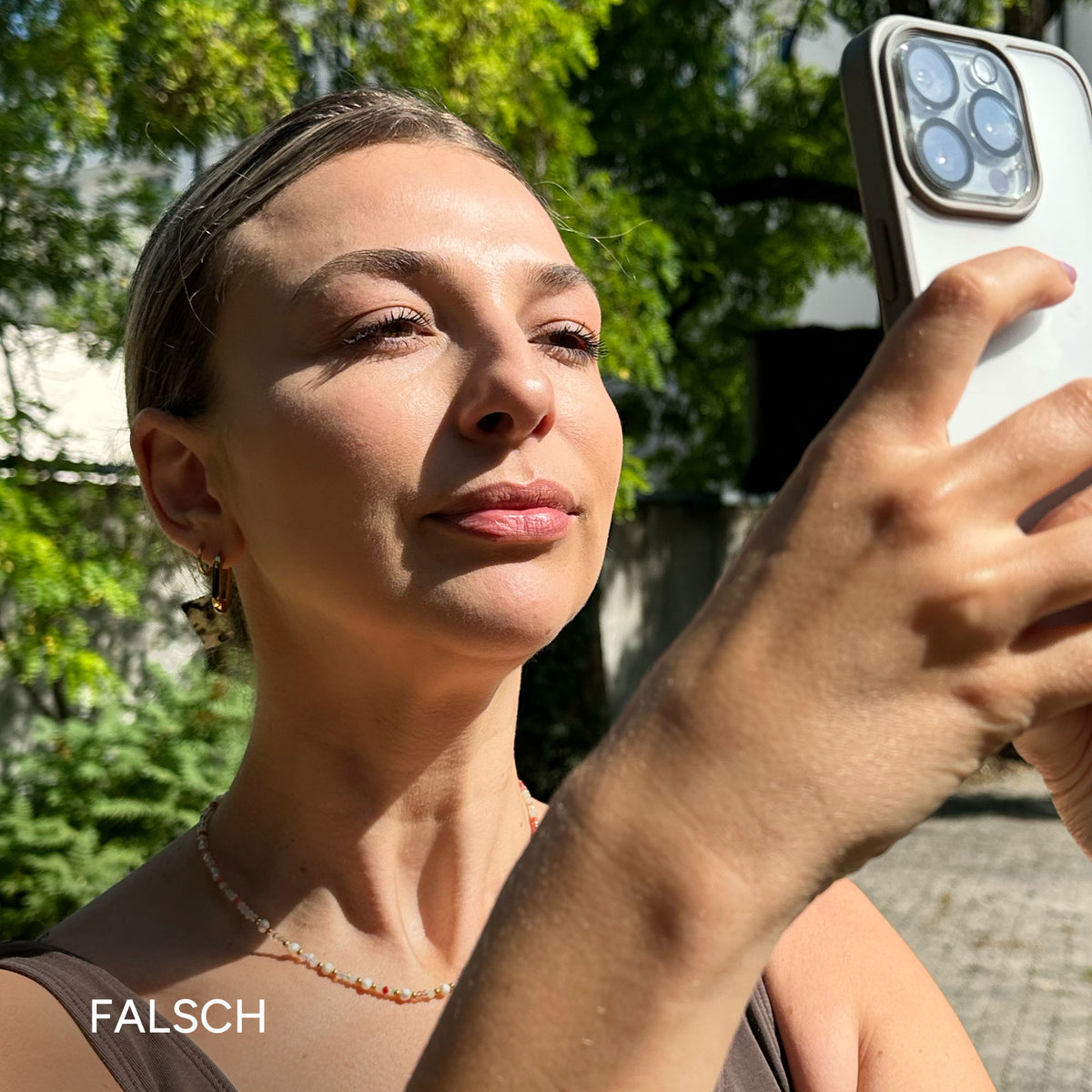 Frau mit kurzem Haar und Schmuck, schaut auf ihr Smartphone im Freien, umgeben von Grün, Lichtspiel auf dem Gesicht.