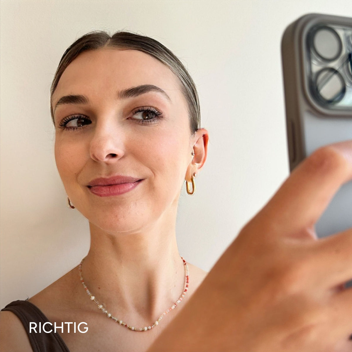 Junge Frau beobachtet sich in ihrem Smartphone, trägt zarte Schmuckstücke und hat ein frisches, natürliches Make-up.