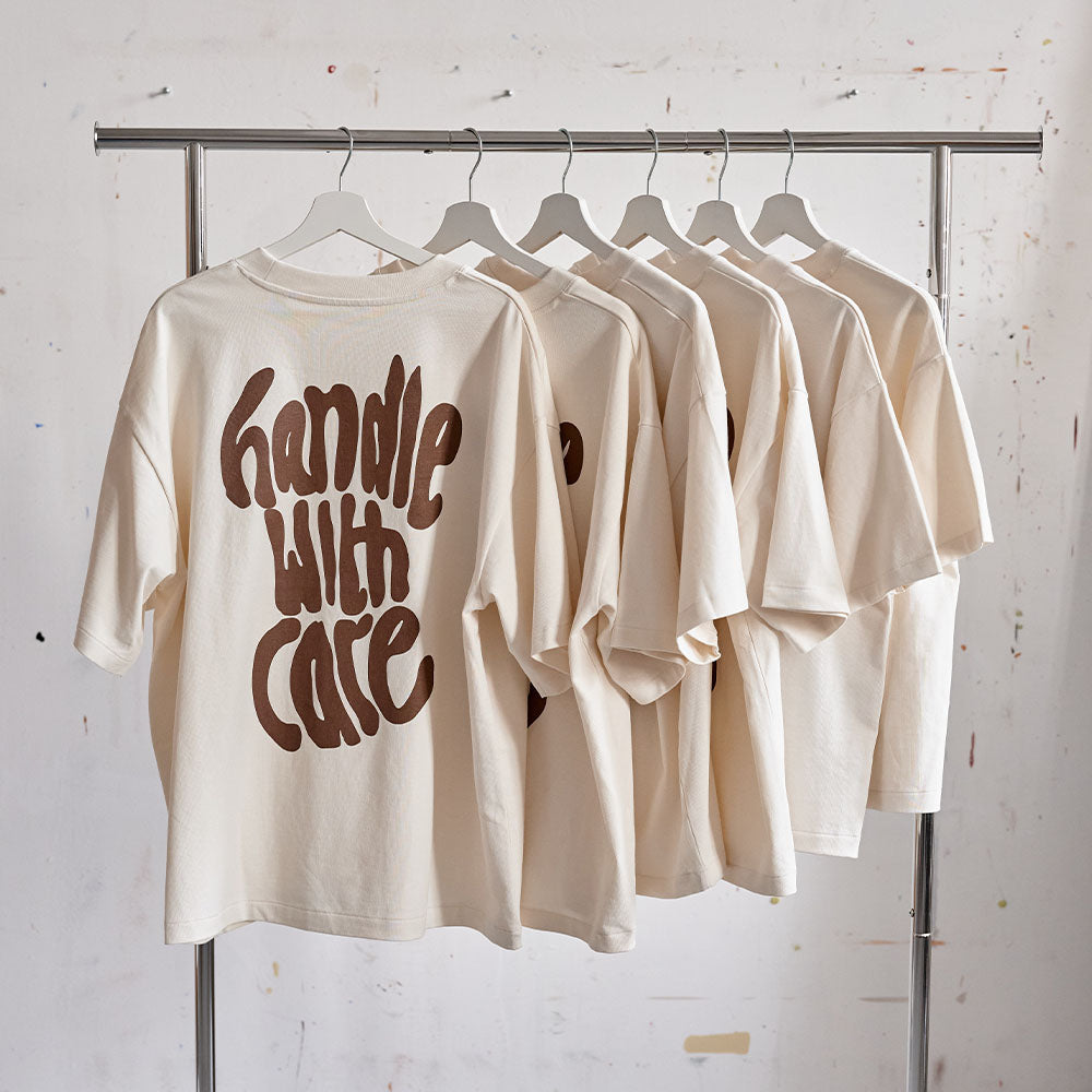 Sechs helle T-Shirts mit dem Aufdruck 'handle with care' hängen auf einer silbernen Kleiderstange.