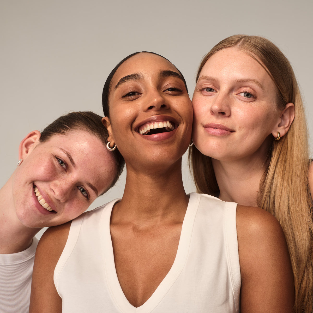 Drei Frauen lächeln zusammen, diverse Hauttöne und Frisuren, warmes, einladendes Ambiente.