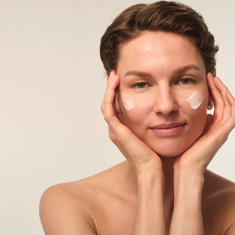 Eine Frau mit kurzem Haar trägt eine Creme auf ihre Wangen auf. Sie lächelt sanft in die Kamera.