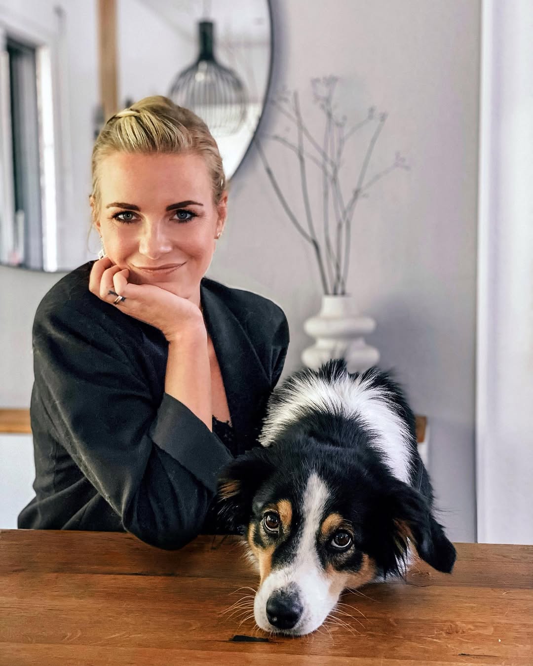 Eine Frau mit blonden Haaren sitzt lächelnd an einem Holztisch. Ihr Hund liegt mit dem Kopf auf dem Tisch.
