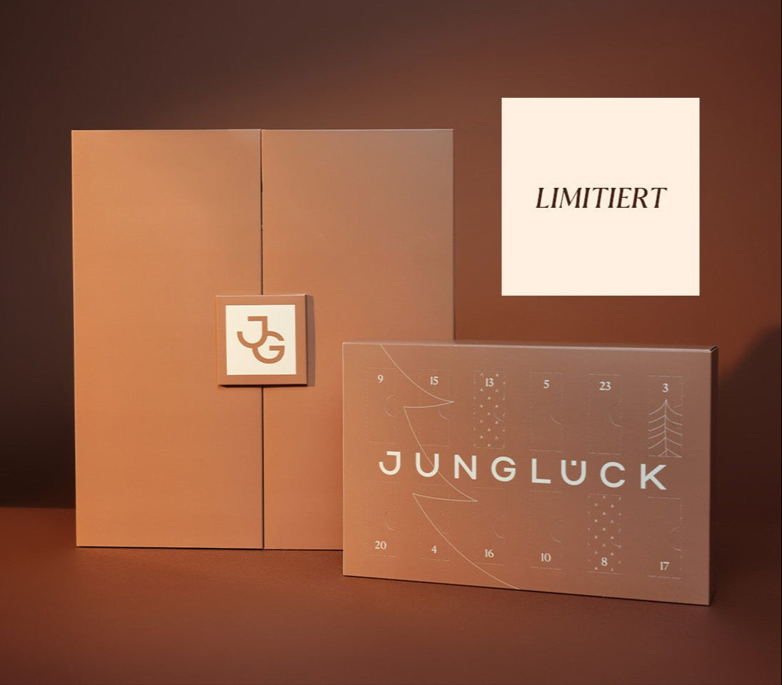 Zwei limitierte Verpackungen von JUNGLÜCK in sanften Erdtönen, eine mit eingestanztem Logo, die andere mit