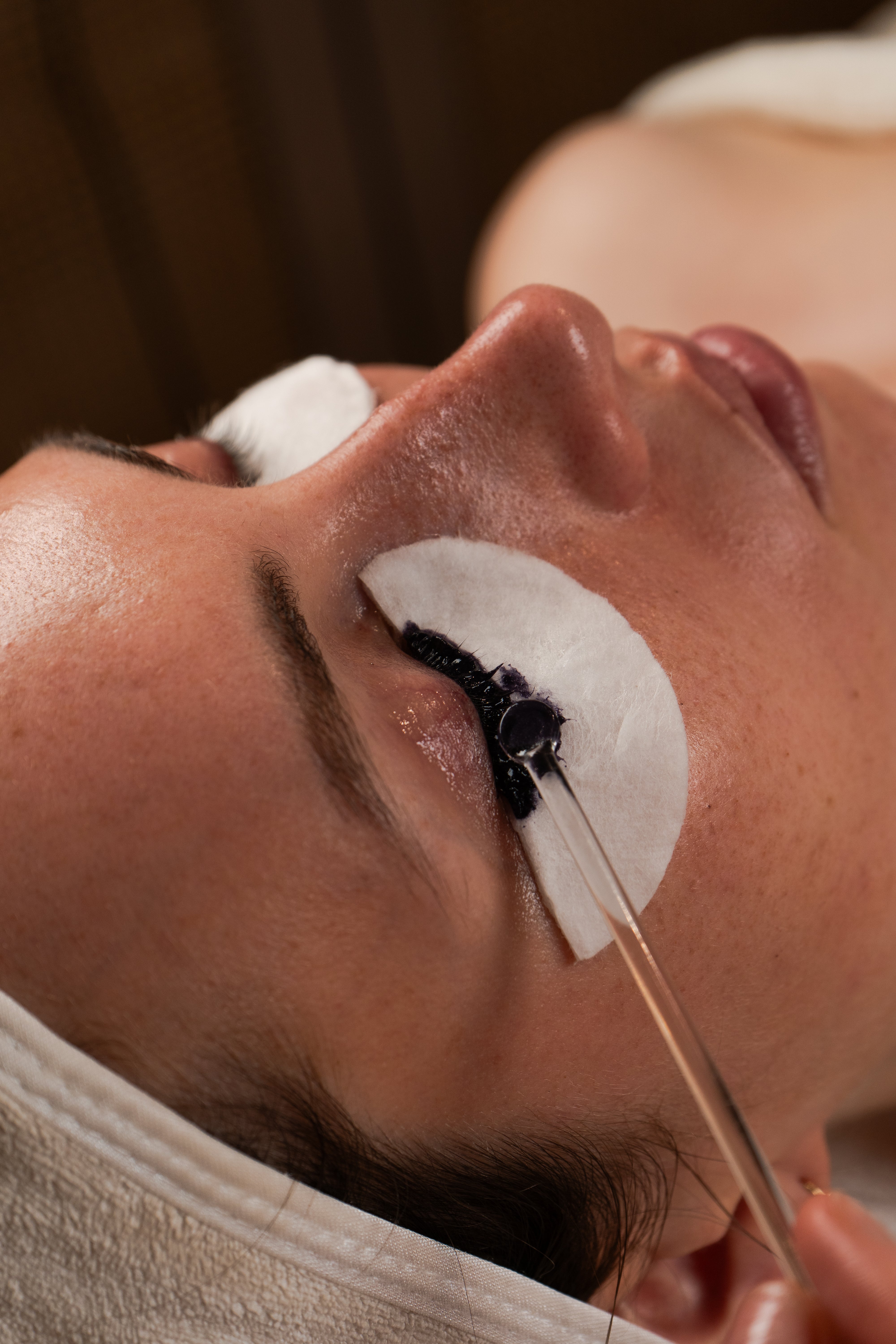 Eine Frau erhält eine Behandlung an den Augenbrauen und Wimpern, dabei wird ein schwarzes Produkt mit einer Pipette