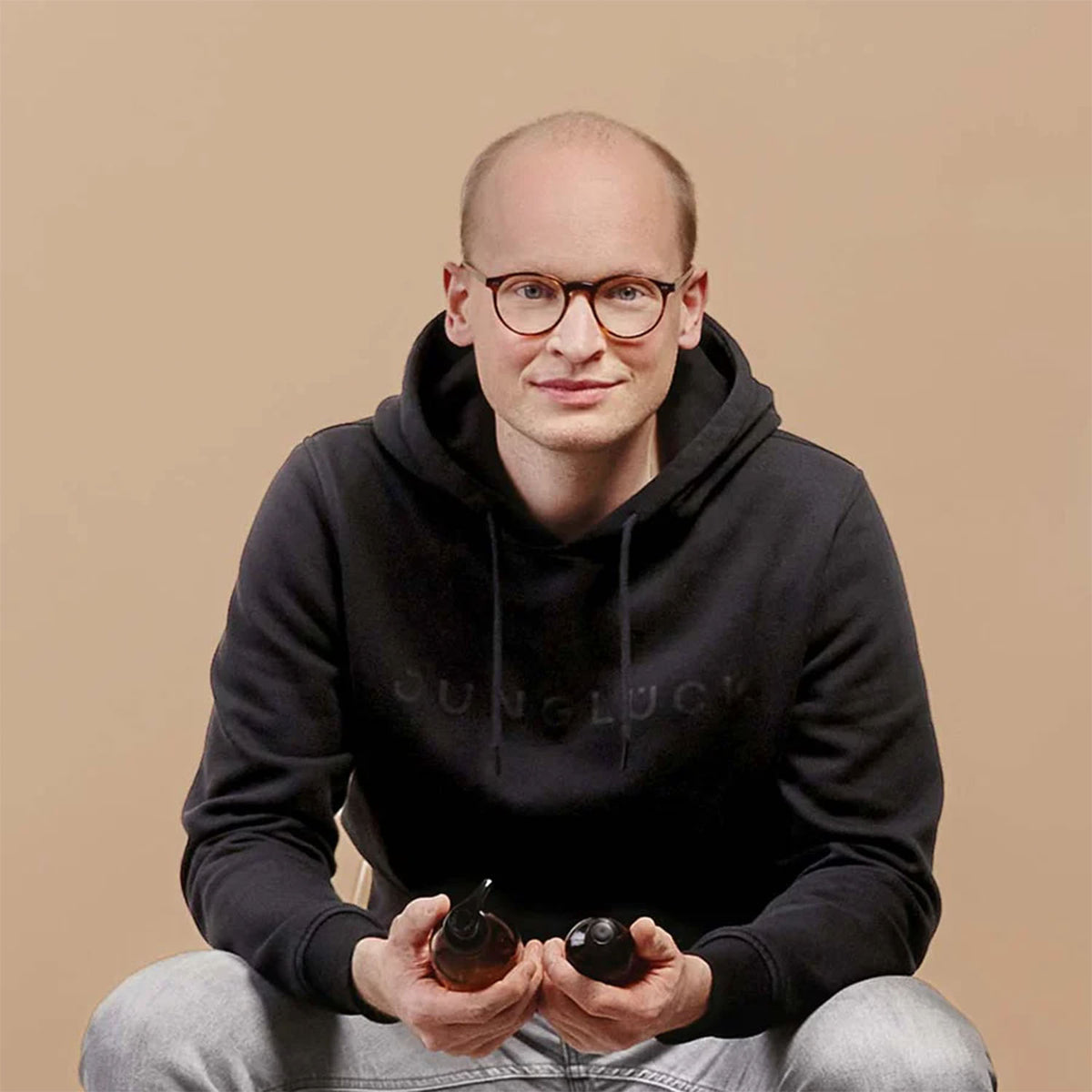Mann mit Brille hält zwei Braunglasflaschen vor einer einfarbigen, hellen Wand. Im Hintergrund ist der Schriftzug JUNGLÜCK