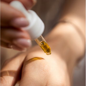 Eine Hand mit zartem Hautton, auf der ein Tropfen goldenes Öl aus einer Pipette fällt.