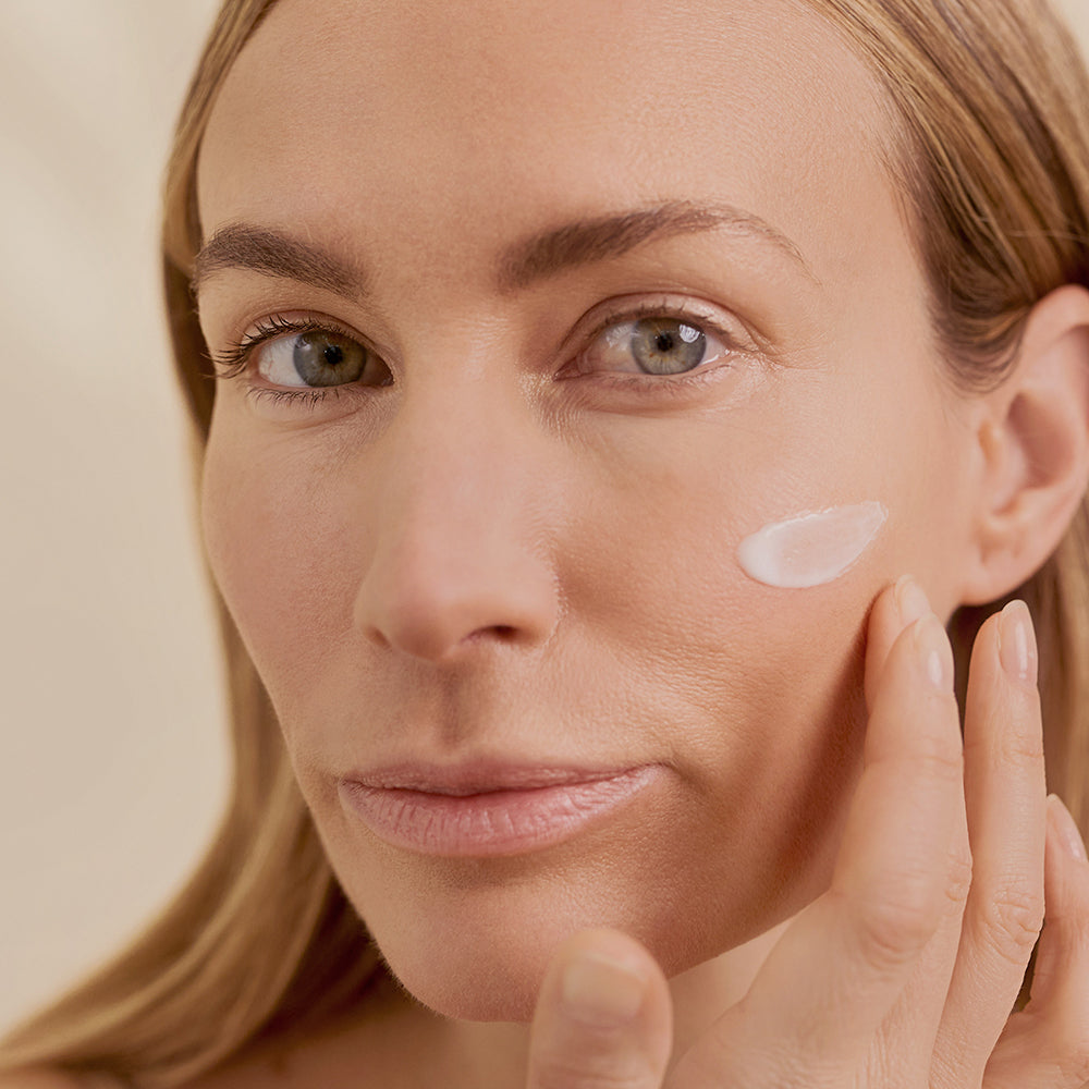 Eine Frau mit leichtem Make-up berührt sanft ihr Gesicht, auf das eine Creme aufgetragen wurde. Die Textur der Creme ist