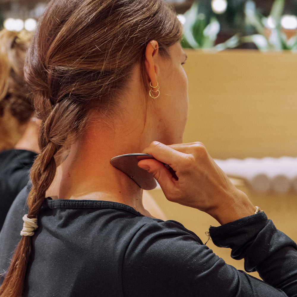 Frau mit geflochtenem Haar führt eine Gesichtsyoga-Übung durch, Anwendung eines Gua Sha Werkzeugs an ihrem Hals.