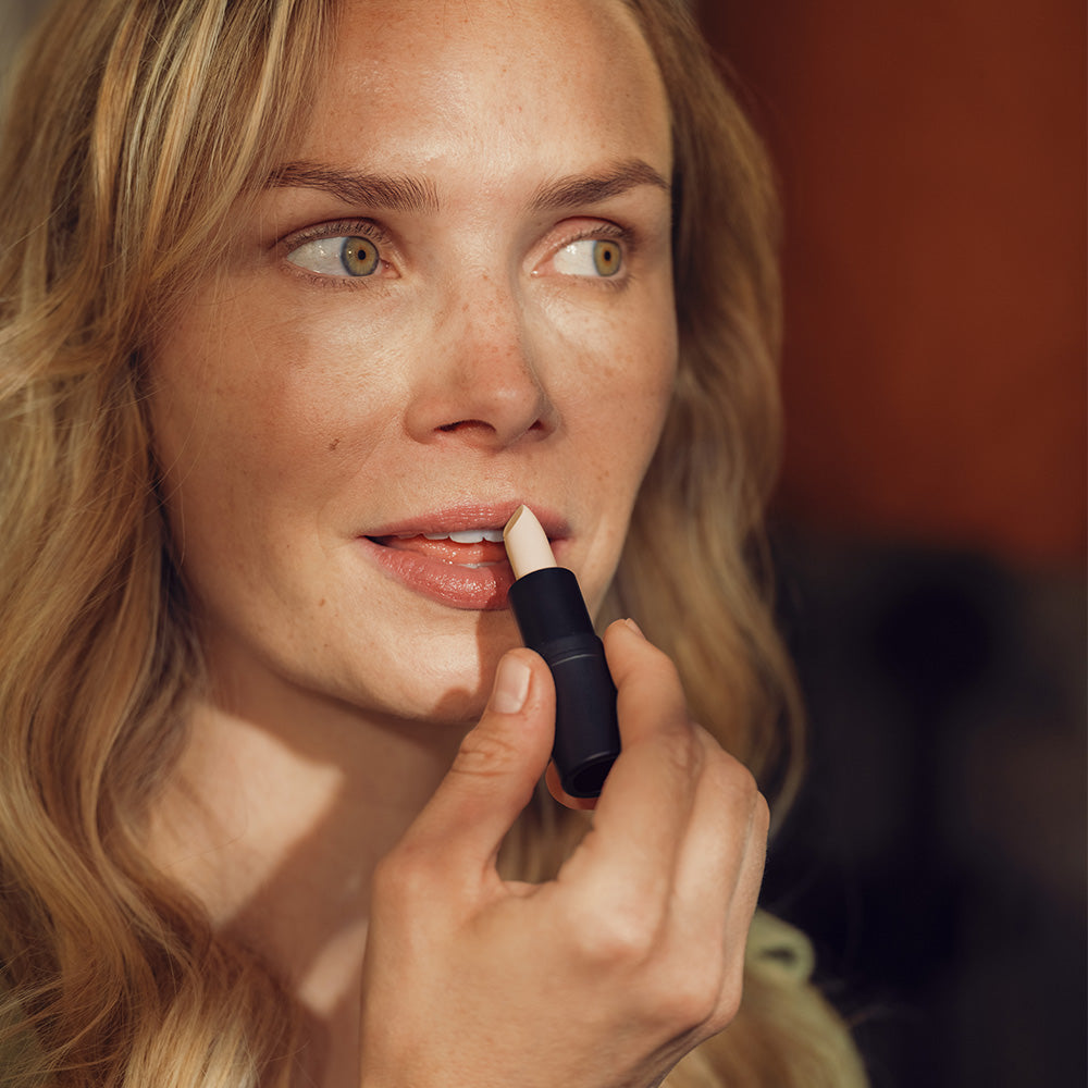 Eine Frau mit lockigem, blondem Haar trägt einen Lippenbalsam auf. Ihr Blick ist sanft und nachdenklich. Das Bild vermittelt