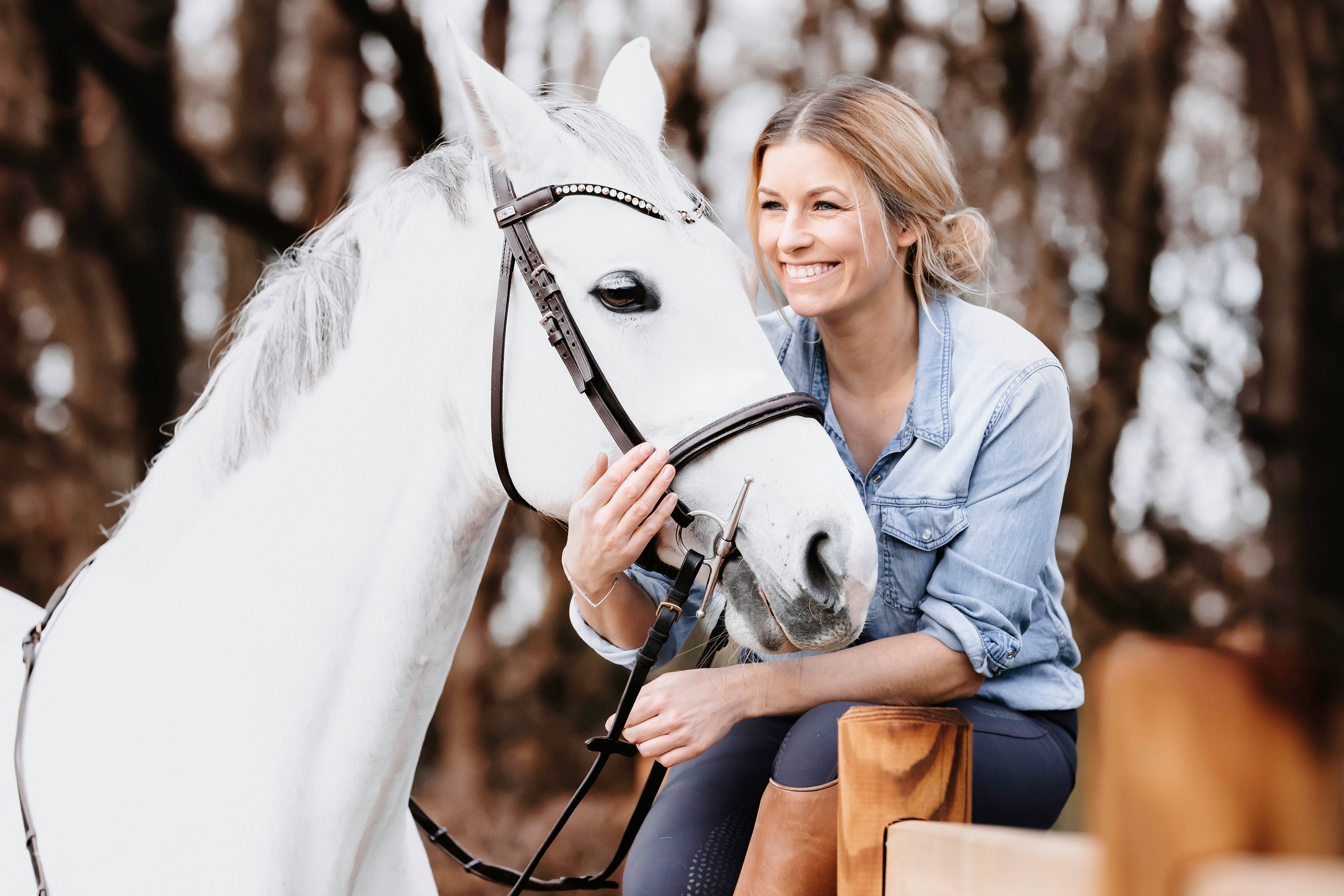 Eine Frau mit blonden, lockigen Haaren sitzt neben einem weißen Pferd und lächelt. Sie trägt ein hellblaues Hemd und braune