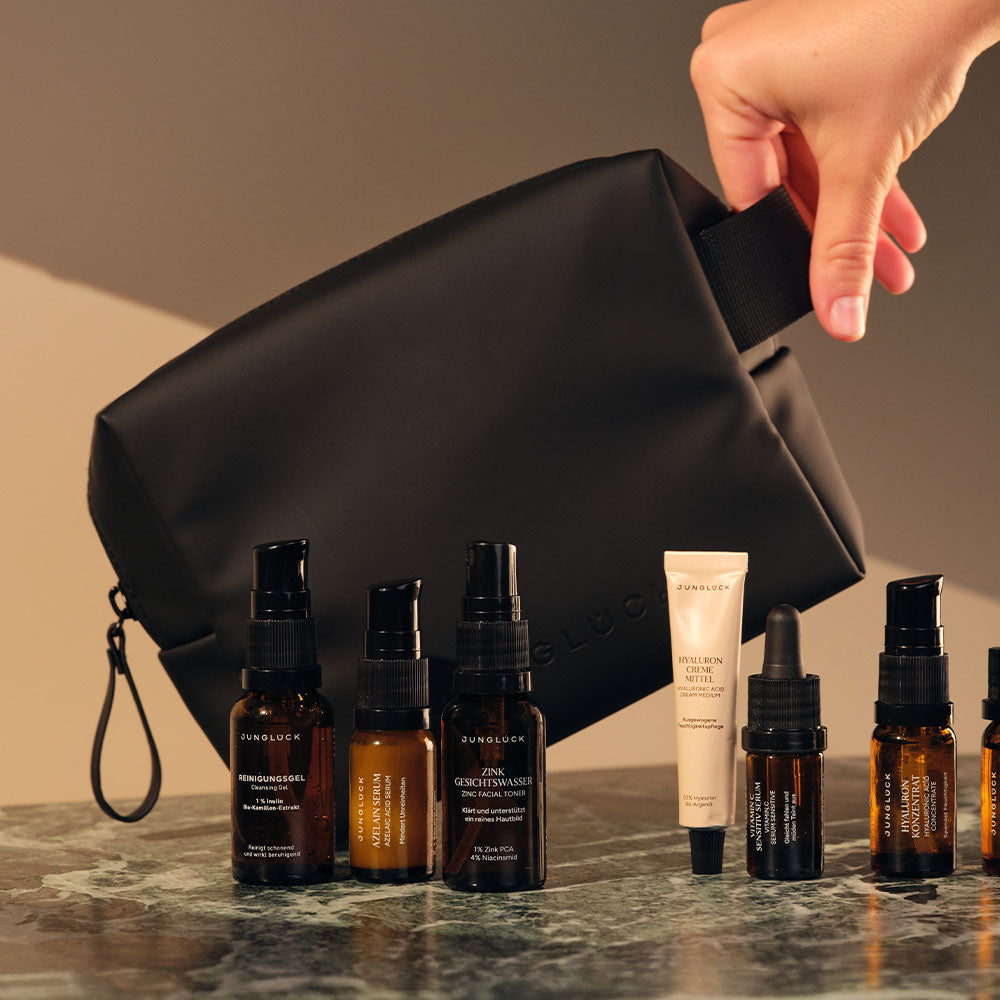 Schwarze Kosmetiktasche wird von Hand gehalten, daneben stehen verschiedene JUNGLÜCK Produkte in Braunglasflaschen.