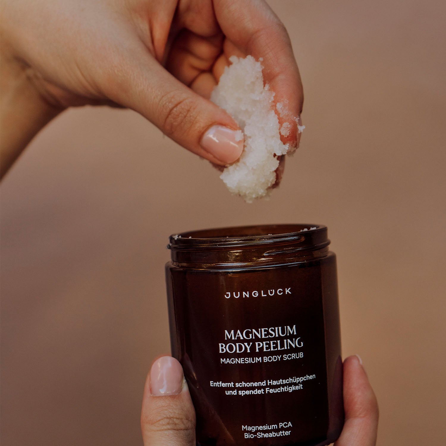Eine Hand hält eine braune Glasflasche mit dem Aufdruck 'Magnesium Body Peeling'. Eine weitere Hand entnimmt mit den Fingern