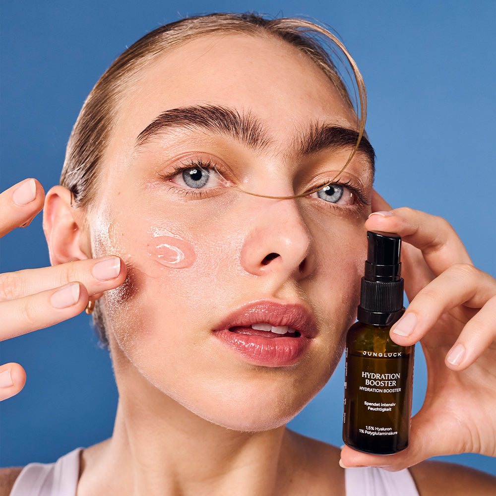 Eine Frau trägt sanft ein Produkt auf ihr Gesicht auf. Sie hält eine Braunglasflasche mit dem Label 'Hydration Booster' in