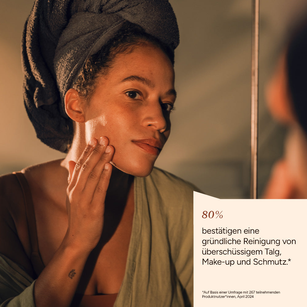 Eine Frau mit einem Handtuch im Haar, die sanft ein Reinigungsgel auf ihrem Gesicht anwendet. Spiegelsituation.