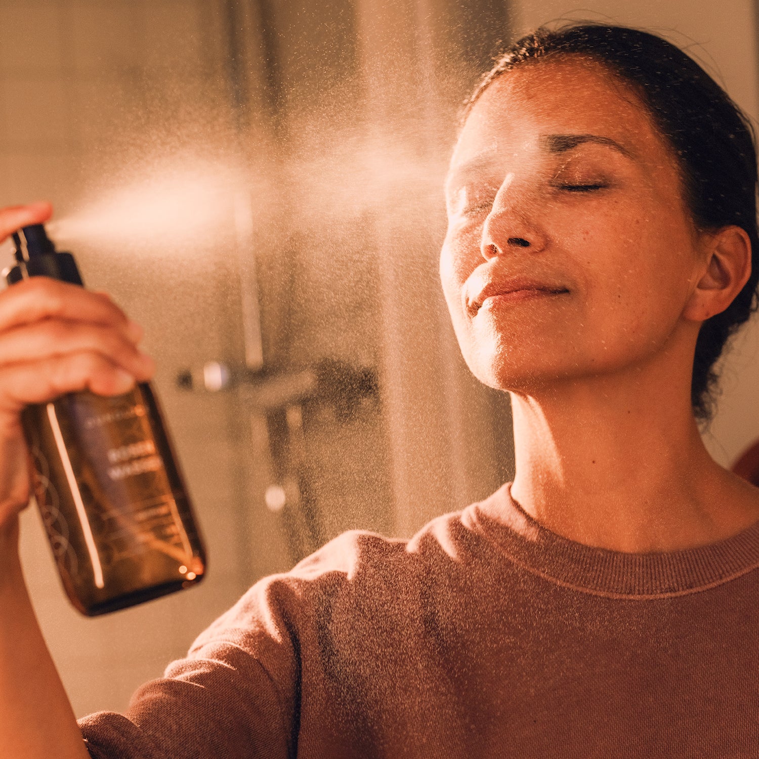Frau sprüht Rosenwasser aus einer Braunglasflasche auf ihr Gesicht, in einem hellen Badezimmer.