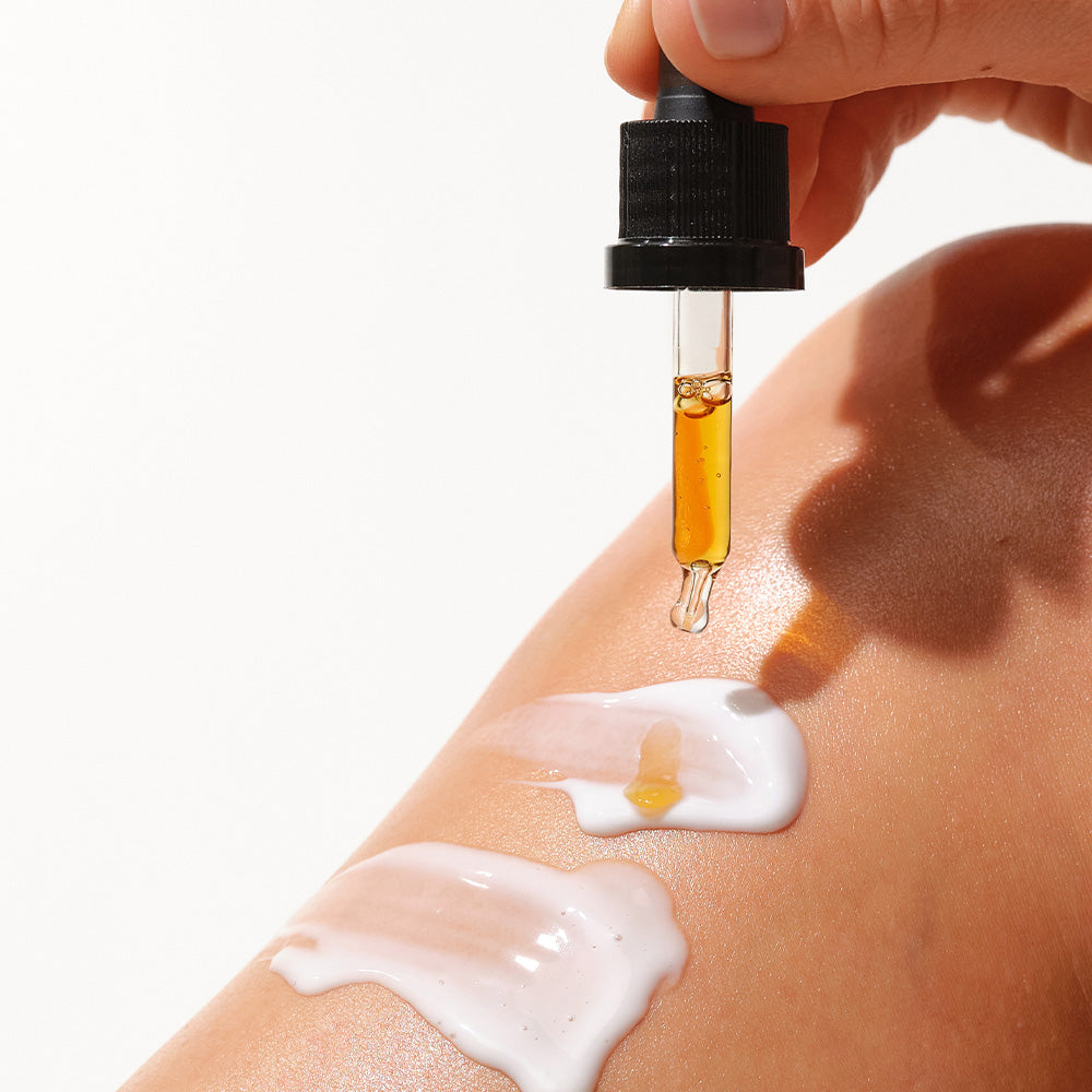 Eine handgehaltene Pipette gibt goldenes Öl auf einen Arm, nebenan ist eine weiße Creme sichtbar.