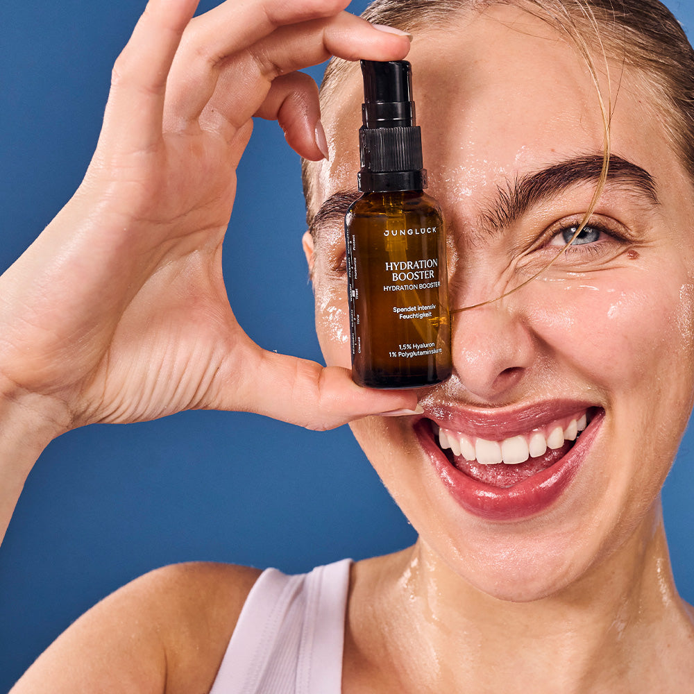 Ein lächelndes Model hält eine Braunglasflasche mit dem Hydration Booster 30ml vor einem blauen Hintergrund. Die Haut des