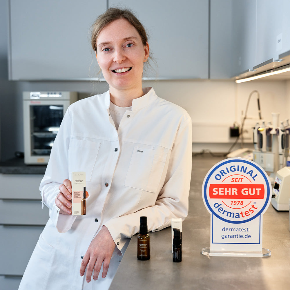 Eine Person in einem weißen Laborkittel hält die Braunglasflasche des Hydration Booster 30ml. Im Hintergrund sind weitere