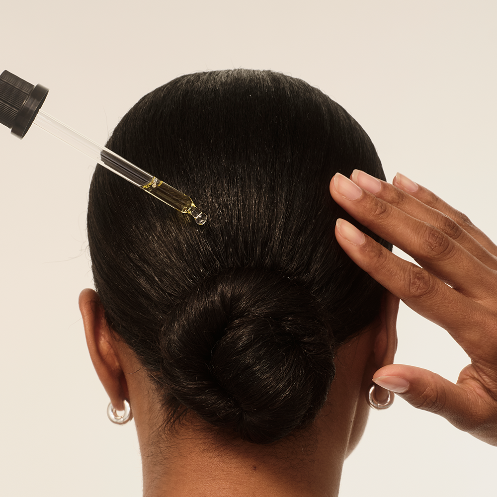Eine Frau mit einem Haarknoten trägt ein Haaröl auf. Die Pipette ist im Haar platziert.