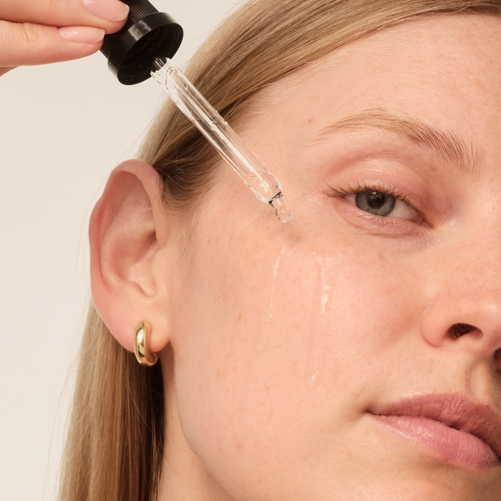 Eine Person trägt Gesichtsserum mit einer Pipette auf die Haut auf. Sichtbare Tröpfchen des Serums auf Wange und Kinn.
