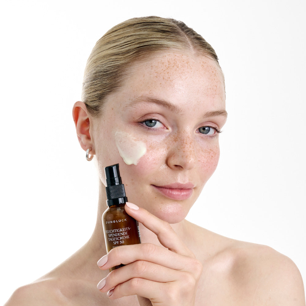 Frau mit Sommersprossen trägt eine Creme auf ihr Gesicht auf. In der Hand hält sie ein Produkt in einer Braunglasflasche.
