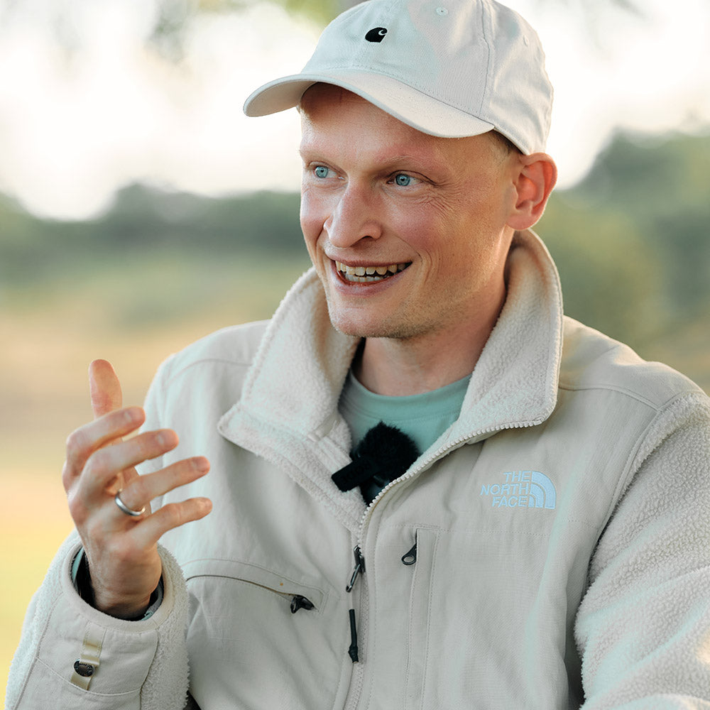 Ein Mann mit Vollbart und kurzen Haaren lächelt während eines Gesprächs. Er trägt eine helle Jacke und eine beige Kappe.