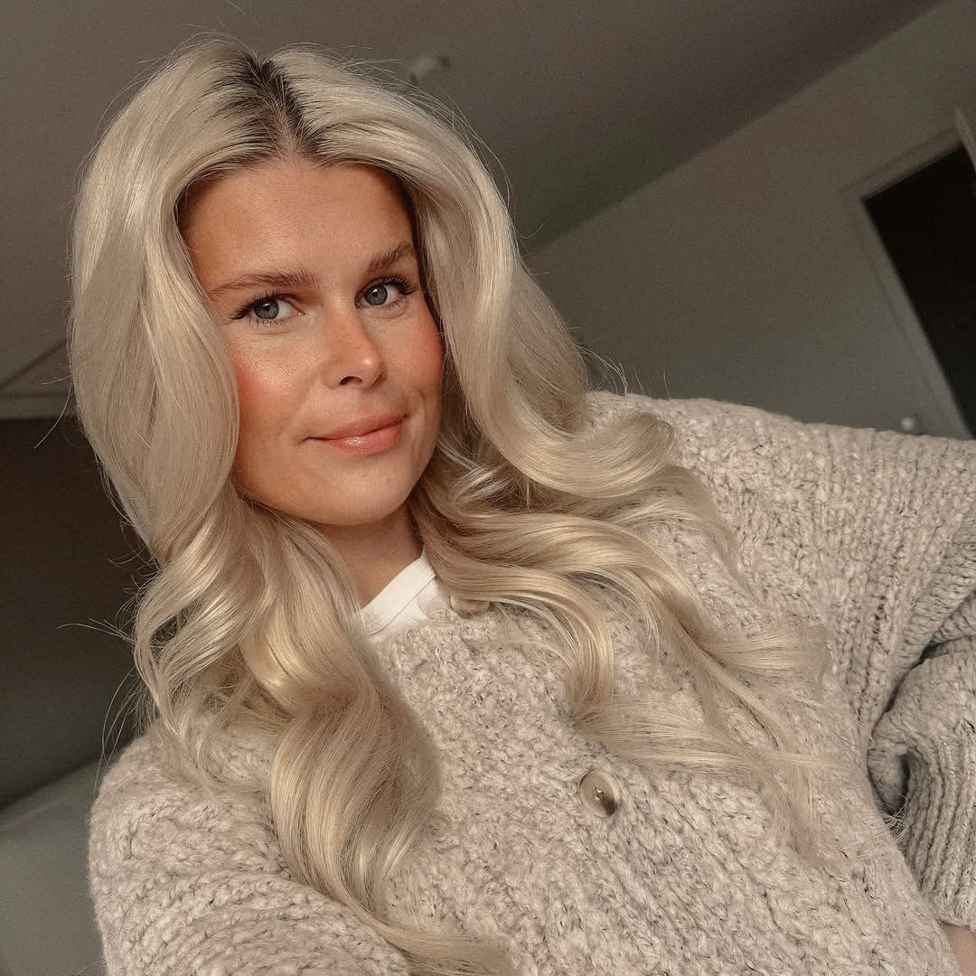 Eine nahaufgenommene Frau mit langen, blonden, welligen Haaren lächelt sanft. Sie trägt einen hellen, strukturierten