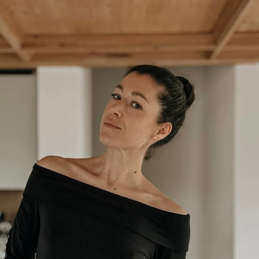 Porträt einer Person mit schulterfreier schwarzer Bluse, in einem hellen Raum mit Holzdecke.