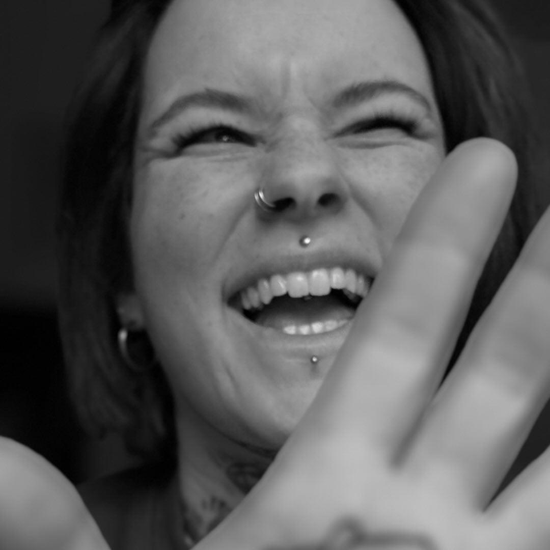 Lächelnde Frau mit Piercings, die die Hand zur Kamera hebt, in schwarz-weiß. Ausdruck von Freude und Unbeschwertheit.