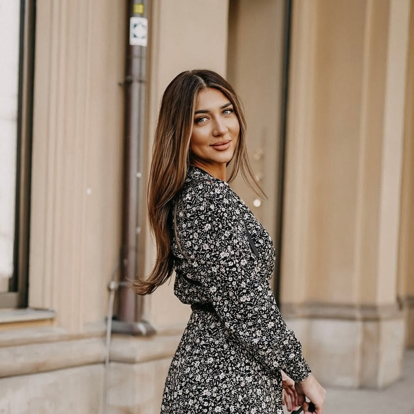 Eine Frau mit langen, braunen Haaren trägt ein florales, schwarzes Kleid und schaut zurück. Der Hintergrund zeigt eine