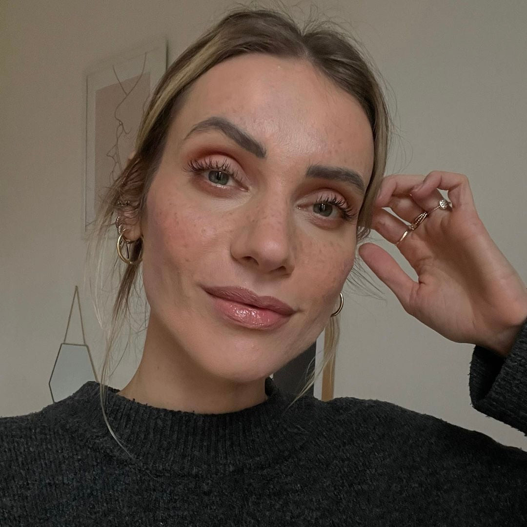 Frau mit natürlichem Make-up, die in die Kamera lächelt, trägt einen dunkelgrauen Pullover und hat lockige Haare.