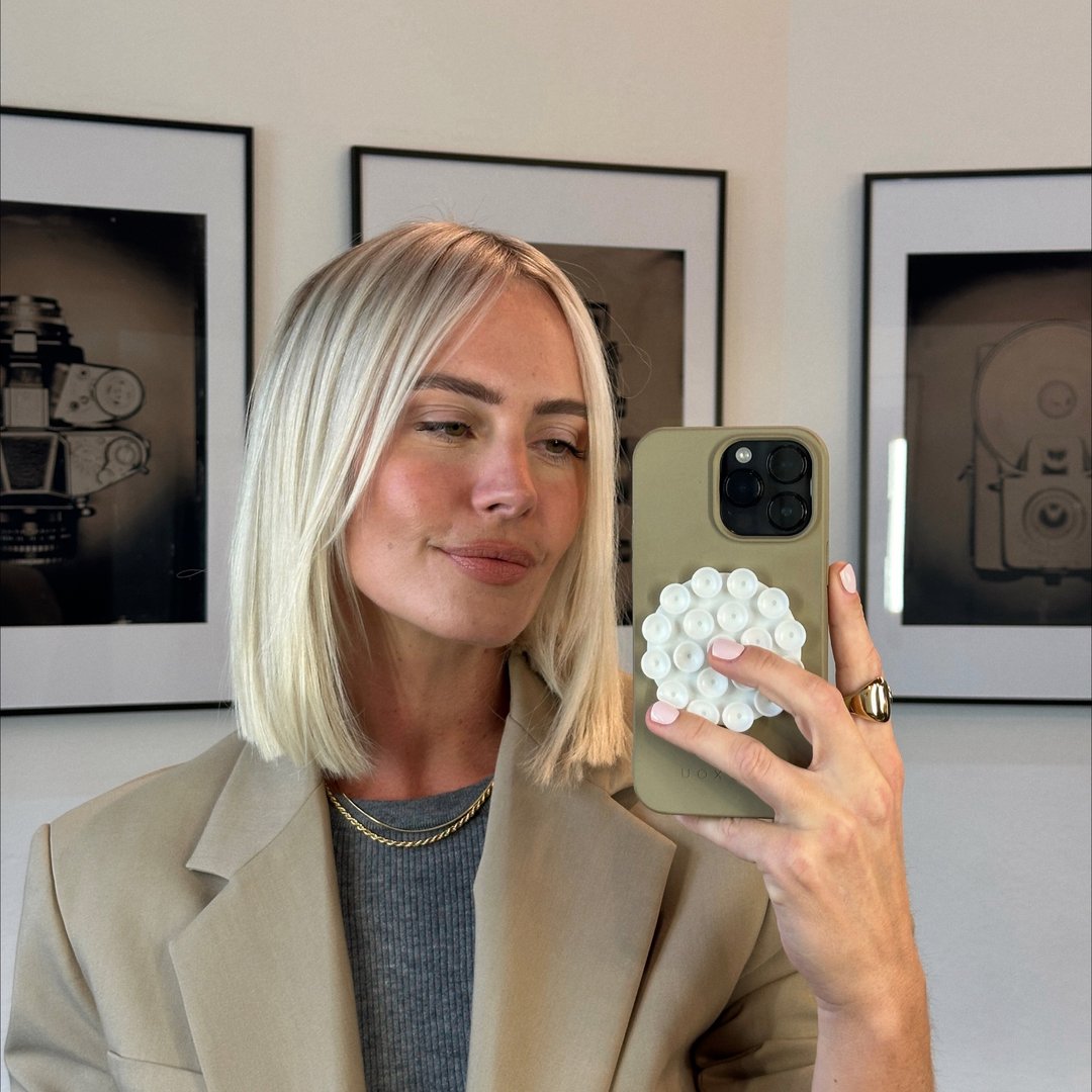 Frau mit blonden Haaren, die in den Spiegel schaut und ein Handy mit einer kreativen Hülle hält. Sie trägt einen Blazer.