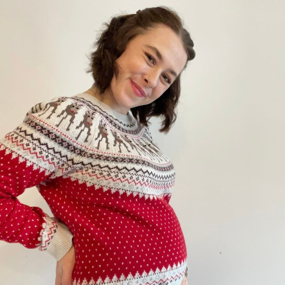 Eine werdende Mutter in einem roten Pullover mit einem winterlichen Muster posiert lächelnd vor einem neutralen Hintergrund.