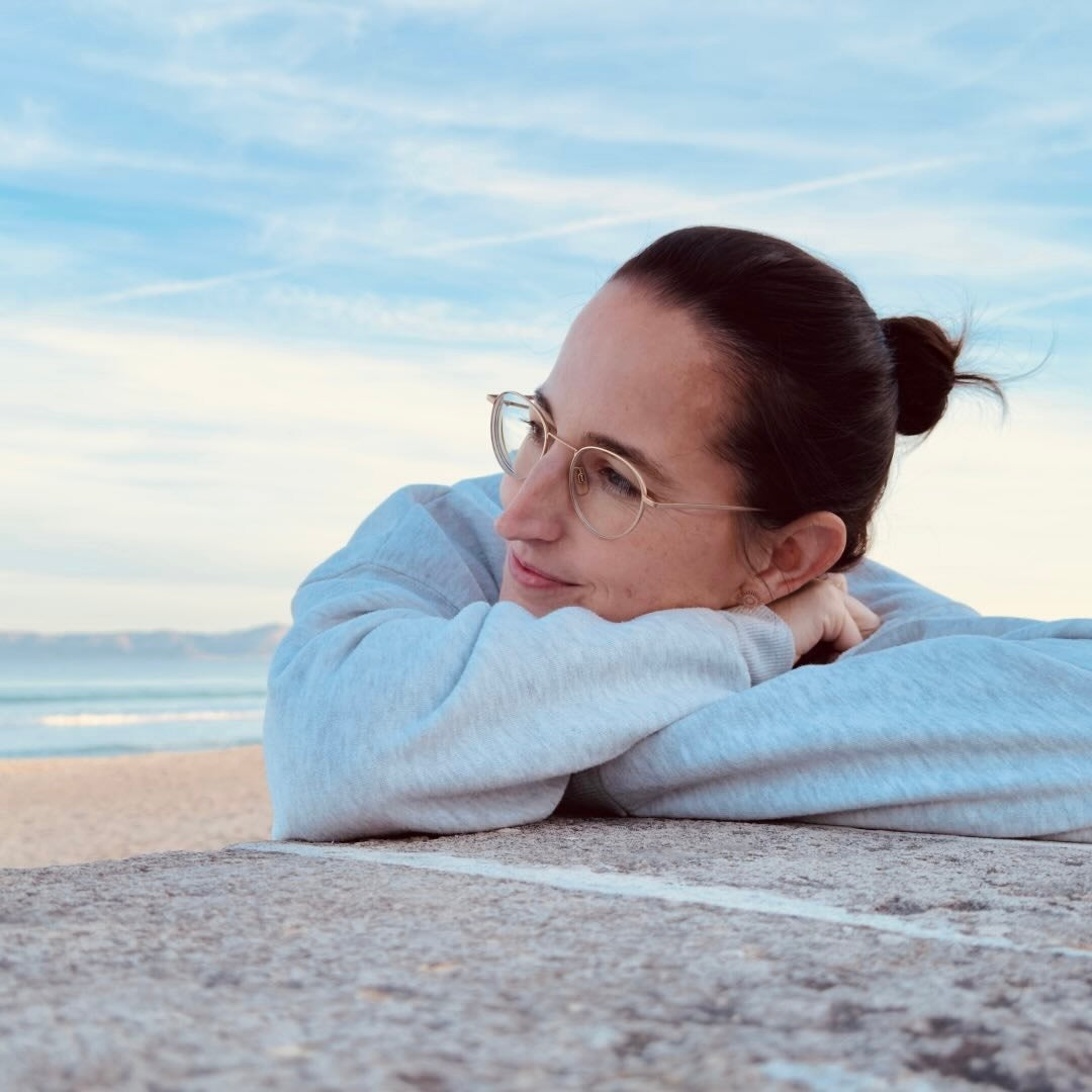 Frau mit Brille und grauem Sweatshirt entspannt am Strand, legt den Kopf auf die Arme, im Hintergrund der Ozean.