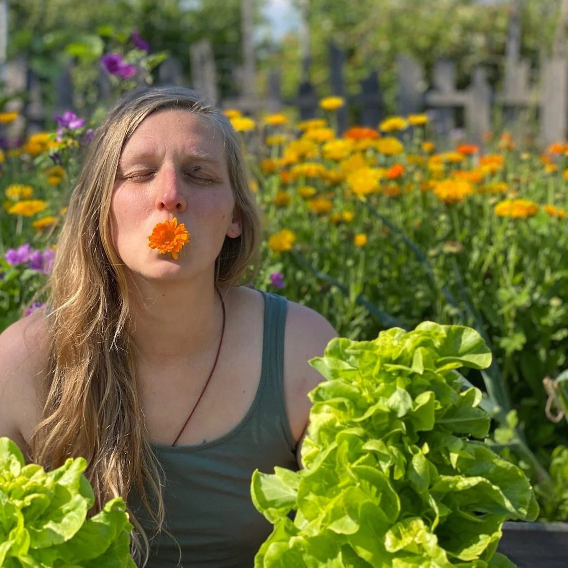Eine Frau sitzt in einem Garten mit bunten Blumen und frischem Salat. Sie hält eine Blüte zwischen den Lippen und lächelt.