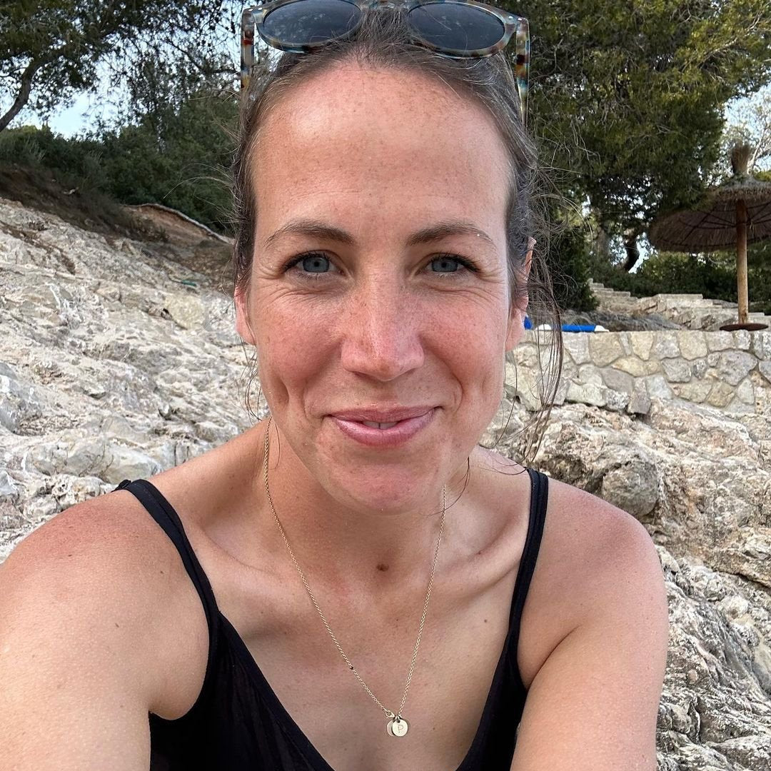 Portrait einer Frau im Freien, lächelnd zwischen Steinen, mit Sonnenschein auf der Haut, trägt eine dezente Kette.