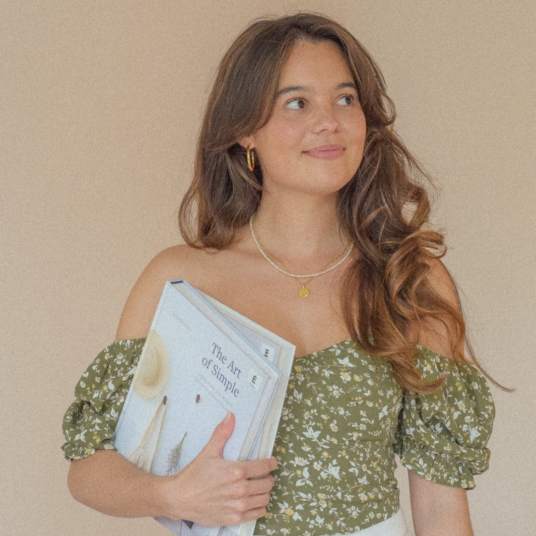 Junge Frau mit lockigem, braunem Haar trägt ein geblümtes Oberteil und hält ein Buch mit dem Titel 'The Art of Simple' in