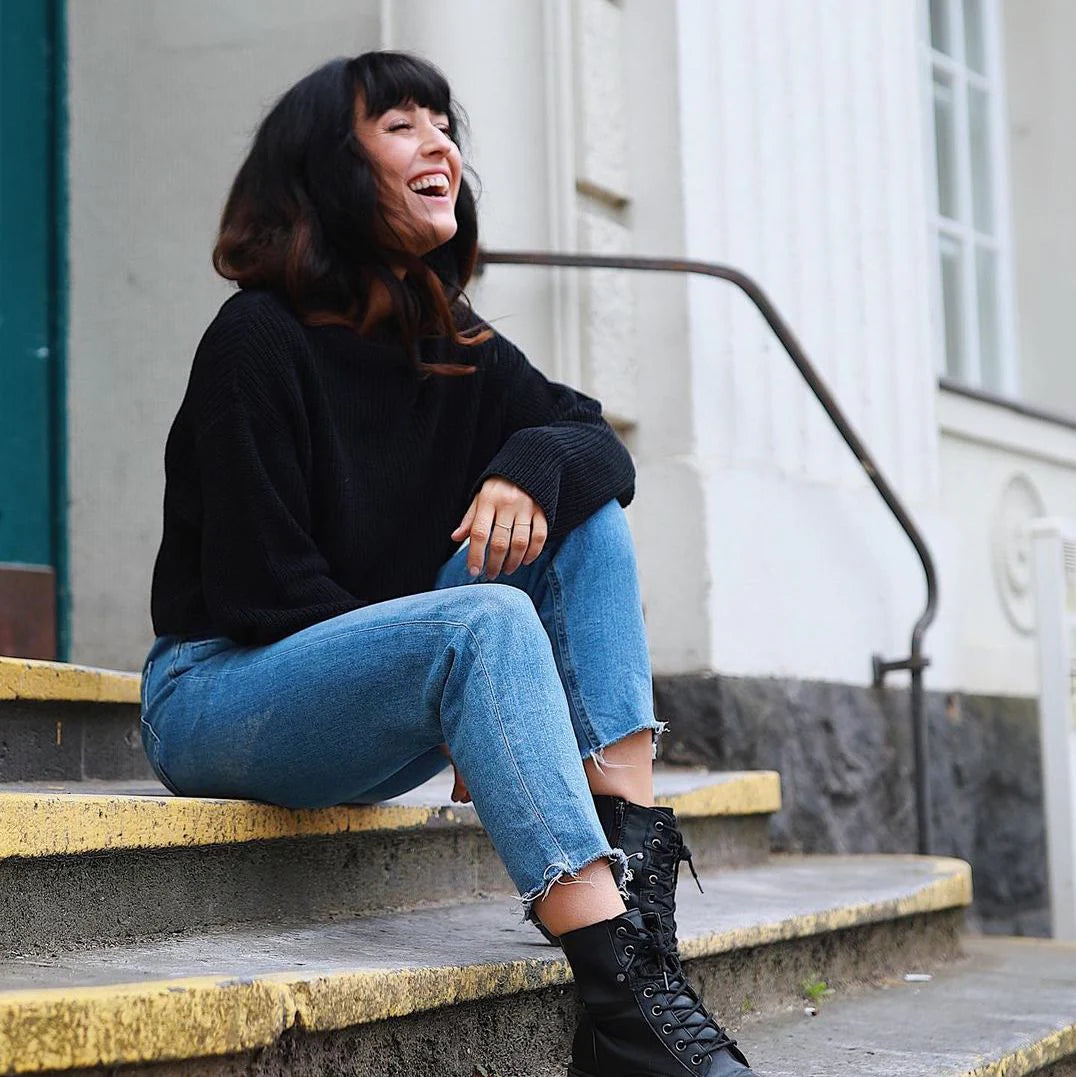 Eine Frau mit schulterlangen, dunklen Haaren, die in einem schwarzen Pullover und blauen Jeans auf Stufen sitzt. Sie lacht