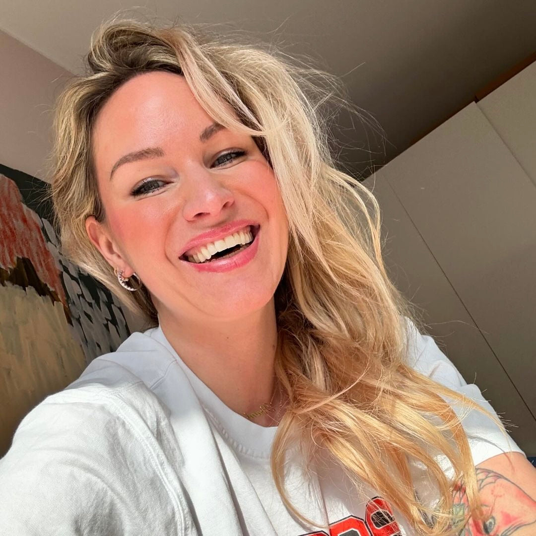 Eine lächelnde Frau mit langer, lockiger blonder Haarpracht trägt ein schlichtes weißes T-Shirt. Der Hintergrund ist