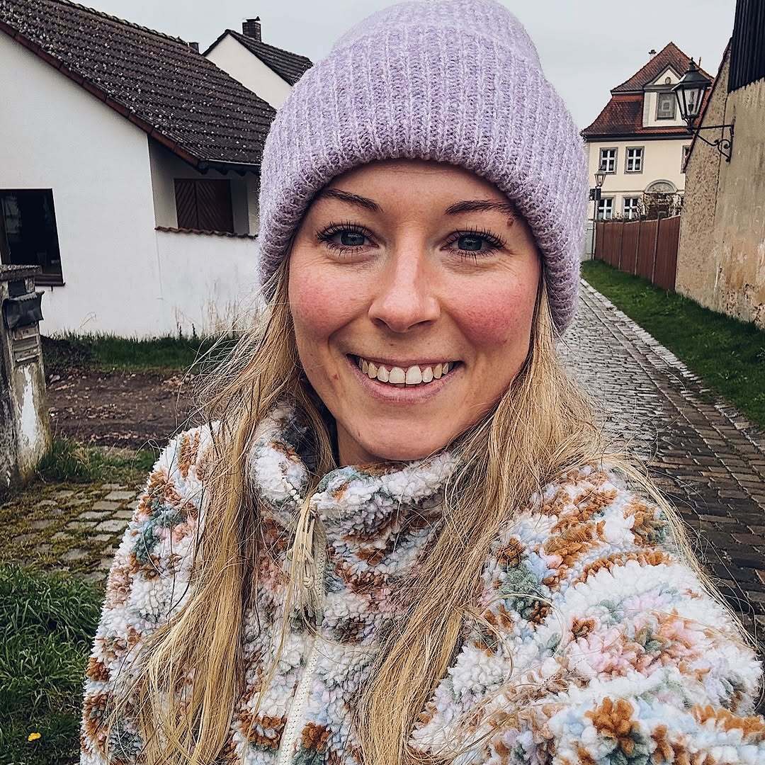 Junge Frau mit einem lila Mütze und einem gemusterten Fleecejacke steht auf einem gepflasterten Weg, umgeben von Häusern.